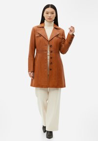 Brun läderrock med notch-krage, knäppning framtill och långa ärmar. Kombinerad med en beige polotröja och vita vida byxor.