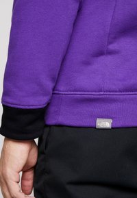 Lila sweatshirt med svarta manschetter, utrustad med en etikett från The North Face. Tyget verkar ha en textur och är designat för komfort.