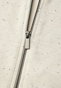 Chaqueta cremosa clara con cierre de cremallera y motas negras, con una cremallera metálica y textura de tela suave. Vista detallada en primer plano del cierre.