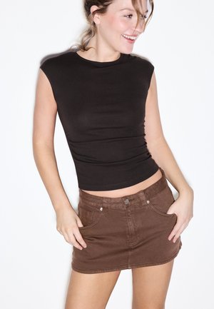 Ung kvinde smiler, iført en ærmeløs sort top og en brun denim mininederdel, står med hænderne i frontlommerne.