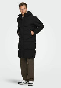 Zwarte gewatteerde jas met capuchon, voorzien van een rits aan de voorkant, drukknoopsluiting, zijzakken en een knielange snit. Gedragen over bruine broek en sneakers.