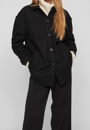 Kobieta ubrana w czarny płaszcz na guziki na biały sweter z golfem i czarne szerokie spodnie, ręce w kieszeniach płaszcza.