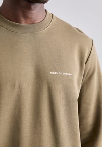 Khaki sweatshirt s kulatým výstřihem, který má jemnou texturu a bílé logo "TIGER OF SWEDEN" na hrudi. Klasický a ležérní design.