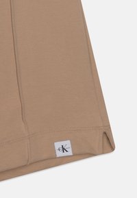 Béžový top z elastické látky s kulatým lemem a bílou etiketou s logem "Calvin Klein Jeans" na dolní levé straně.