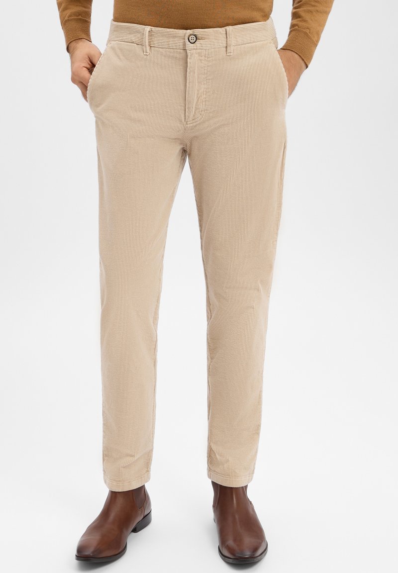 Beżowe spodnie sztruksowe o pionowej, prążkowanej fakturze, wąskim kroju, zapięciem na guziki i bocznymi kieszeniami, w połączeniu z brązowymi skórzanymi butami.