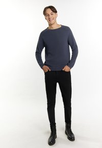 Maglione texturizzato blu navy con scollatura rotonda, abbinato a pantaloni slim-fit neri e stivali neri con lacci. Il modello sta in posizione rilassata con le mani in tasca.