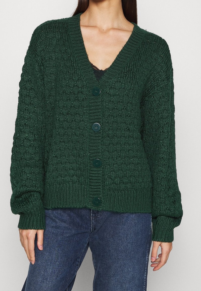 Femme portant un cardigan en maille texturée vert foncé à boutons, par-dessus un haut noir et un jean bleu, debout les bras détendus le long du corps.