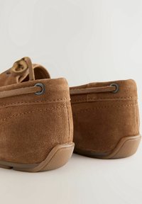 Chaussures mocassins en daim marron avec des lacets en cuir passant par des œillets métalliques, vues de dos sur un fond neutre.