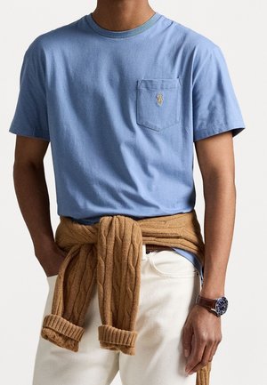 Homme portant un T-shirt bleu clair à manches courtes avec une poche poitrine et un petit logo, un pull tricoté beige noué autour de la taille, un pantalon blanc et une montre-bracelet.