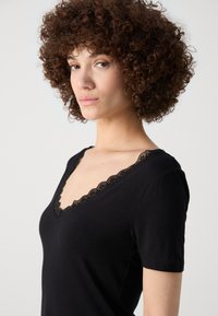 Svart kortermet skjorte med V-hals og delikat blondedetalj. Stoffet ser mykt og glatt ut, noe som fremhever en tettsittende Silhouette.