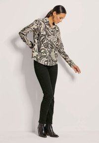 Langärmlige Bluse in Creme mit schwarzem Paisleymuster, mit Kragen und Knopfverschluss. Kombiniert mit eng anliegenden schwarzen Jeans und schwarzen Knöchelstiefeln.