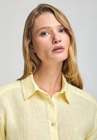 Ljusgul linneskjorta med nedknäppbar krage, en bröstficka och subtil textur. Har ljusa knappar och minimal detaljering.