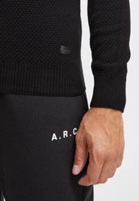 Czarny dzianinowy sweter o teksturowanym wzorze, z prążkowymi mankietami i dolnym wykończeniem oraz małą naszywką z logo z boku. Noszony z czarnymi spodniami dresowymi z białymi napisami.