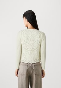 Maglione lavorato a maglia verde chiaro con un motivo in pizzo testurizzato, maniche a costine e un taglio cropped. Indossato sopra pantaloni beige a vita alta.