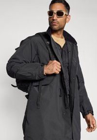 Black, hooded parka with a zip and snap front closure, featuring multiple pockets and a textured fabric. Worn with beige sunglasses.