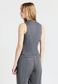 Femme avec des cheveux blonds raides, portant un haut sans manches à rayures grises et un pantalon assorti, tournée vers la gauche sur un fond blanc.