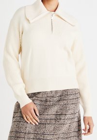 Pull-over en maille crème avec col haut et fermeture éclair. Présente une texture côtelée sur les manches et l'ourlet inférieur, porté avec une jupe marron à motifs.