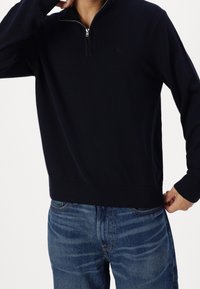 Maglione blu navy con collo mezza cerniera, polsini e orlo a costine; indossato sopra jeans blu. Texture morbida e design minimalista.