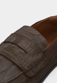 Mocassin en cuir marron foncé avec texture florale embossée et bride à boucle sur le dessus, présentant des détails de couture visibles.