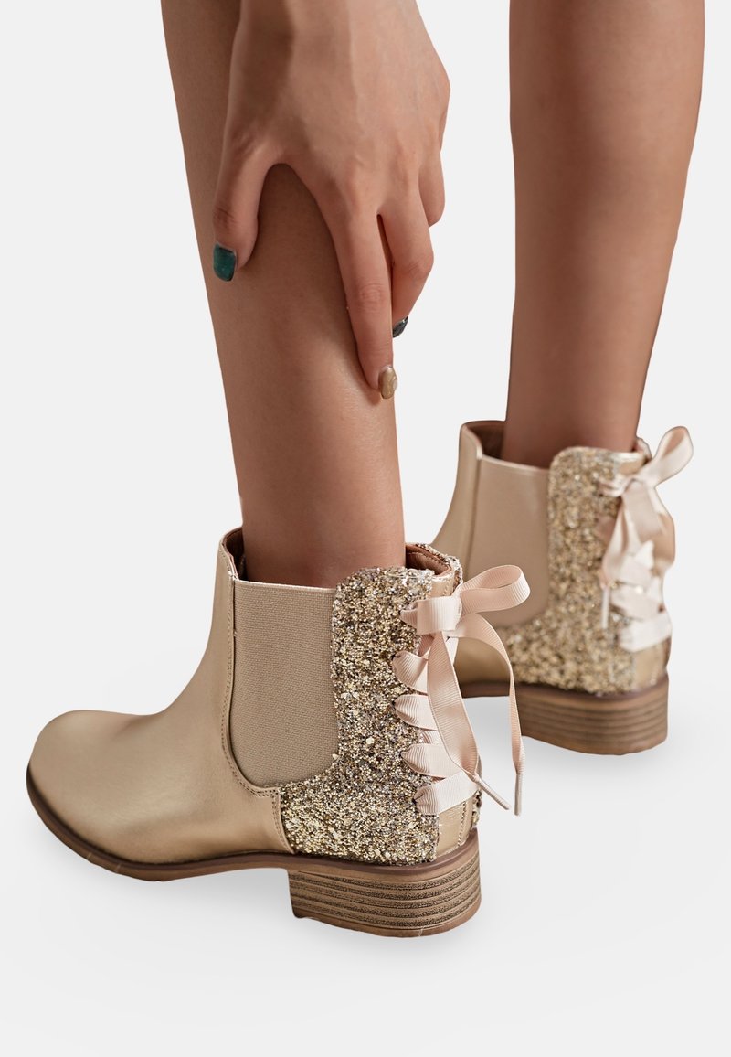 Bottes beiges à cheville avec des arrière en or scintillant et des nœuds en satin, portées sur des jambes nues avec une main touchant doucement la jambe droite.