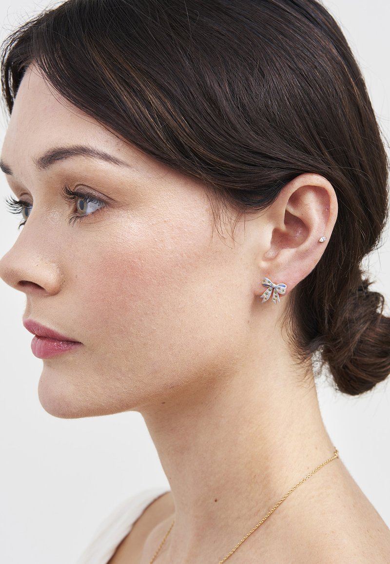 Young woman with dark hair in a low bun wearing a silver bow-shaped earring and a small stud on her right ear against a plain background.