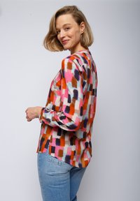 Kolorowa bluzka w wzory z abstrakcyjnym motywem pociągnięć pędzla w odcieniach pomarańczowym, różowym i niebieskim, z długimi rękawami i luźnym krojem, zestawiona z jasnymi dżinsami.