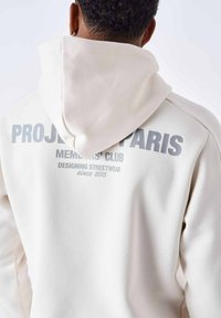 Off-white hoodie met een trekkoord, met grote grijze tekst op de achterkant: "PROJECT PARIS MEMBERS' CLUB" en "DESIGNING STREETWEAR sinds 2015."