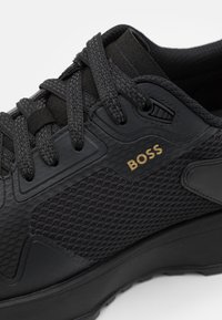Zwarte sportieve schoen met een gestructureerd mesh en gladde synthetische materialen. Veters in het zwart met een gouden "BOSS" logo accent aan de zijkant.