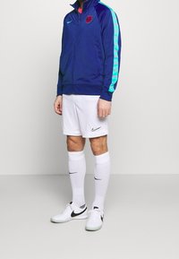 Giacca a vento della marina con strisce laterali turchesi, stemma rosso; pantaloni corti bianchi con logo Nike; calze bianche fino al ginocchio; scarpe da ginnastica bianche e nere.