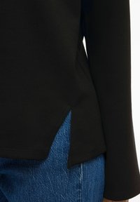 Juodas ilgas rankovės marškinėlis su suapvalintu hemu, turintis šonines skyles. Derinamas su šviesiai mėlynomis džinsais, turinčiais laisvą siluetą ir lygią tekstūrą.