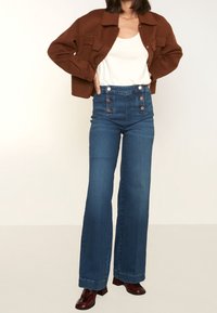 Korkeavyöhykkeiset siniset farkut, joissa on levenevä lahje ja etupainiketta, yhdistettynä ruskeaan lyhytvartiseen takkiin ja valkoiseen toppiin.