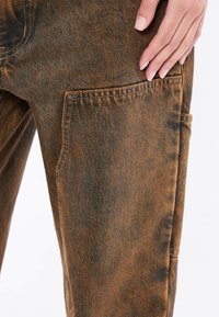 Bruine denim broek met een verouderde textuur, zichtbare stiksels en een zijzak met een drukknoopdetail.