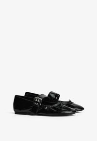 Schwarze Lacklederschuhe mit spitzem Zehenbereich, Rüschensaum und einem Riemen mit dekorativen Beschlägen. Flache Sohle.