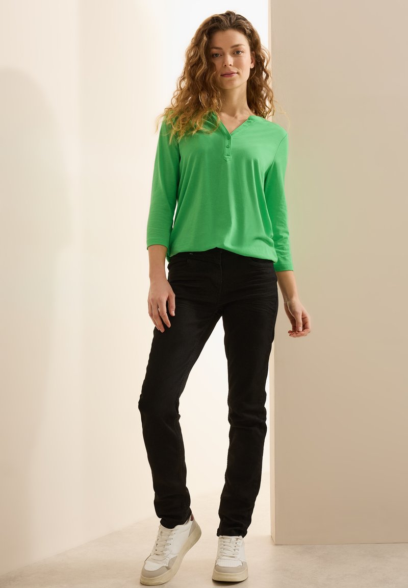 Grön långärmad henley skjorta, svarta tight jeans och vita sneakers. Skjortan har en V-ringning och knappar, med en mjuk tygstruktur.