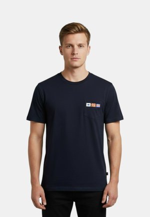 Giovane uomo con capelli biondi corti che indossa una maglietta blu navy con tre piccole bandiere colorate sulla tasca sinistra del petto.