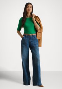 Zelený priliehavý top, modré džínsy s vysokým pásom a širokými nohavicami s veľkými vreckami, hnedá kožená bunda prehodená cez rameno, zlaté kovové doplnky.