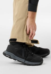 Scarpe da trekking impermeabili nere con accenti grigi, dotate di una parte superiore texturizzata e suola robusta, abbinate a pantaloni beige fissati con Velcro.