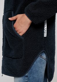 Marinblå fleecejacka med texturerad yta och sidoficka. Har kontrasterande kantband längs fickor och nederkant, samt en lätt rundad nederdel.