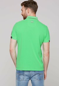 Zielona koszulka polo z klasycznym kołnierzykiem, krótkimi rękawami i fakturowanym materiałem. Posiada naszywkę z logo na lewym rękawie i kontrastowe szwy.