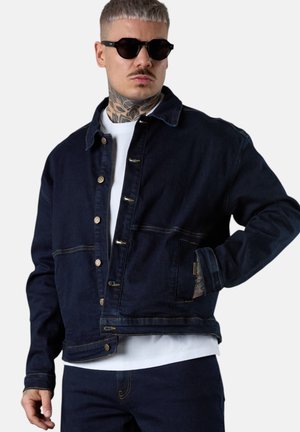 Mann mit Nackentattoo, der schwarze Sonnenbrille, eine dunkle Jeansjacke über einem weißen Hemd und dazu passende dunkle Jeanshosen trägt.