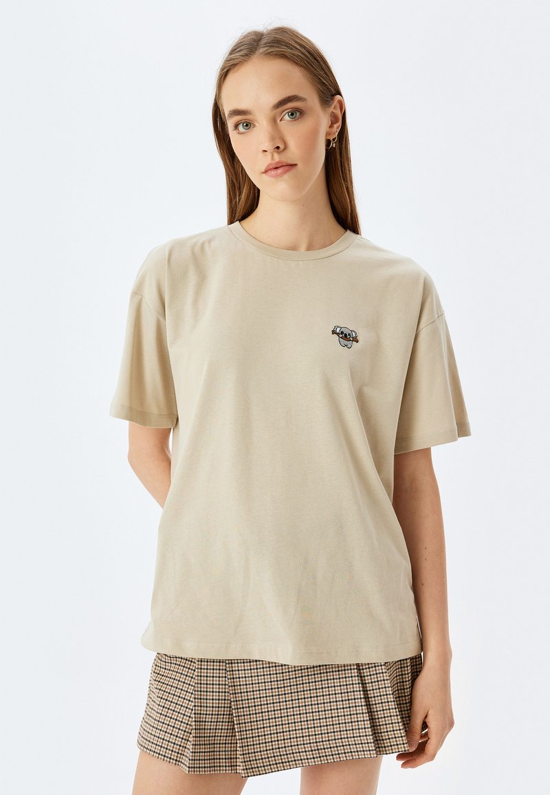 Beige bomulds T-shirt med korte ærmer og rund halsudskæring. Har et lille grafisk motiv på brystet. Sammen med et ternet nederdel.