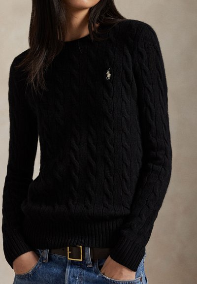 Mujer que lleva un suéter de punto con trenzas negro con un pequeño logo bordado en el pecho, vaqueros azules y un cinturón marrón con hebilla dorada.