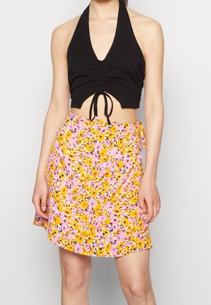 Kobieta ubrana w czarny, bezrękawnikowy top z wiązaniem na szyi i różowo-żółtą kwiecistą spódnicę na zakładkę, stojąca na jednolitym tle.