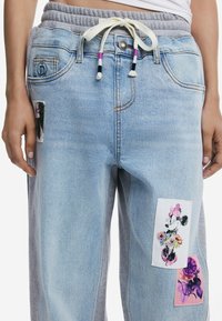 Lichtblauwe denim joggers met een grijze tailleband, voorzien van kleurrijke stoffen patches: een bloemmotief en een karakterprint aan de voorkant.