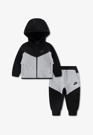 Dwuczęściowy dres Nike składający się z czarno-szarej bluzy z kapturem na zamek błyskawiczny z wzorem w kształcie szewronu oraz dopasowanych spodni dresowych o zwężanych nogawkach.