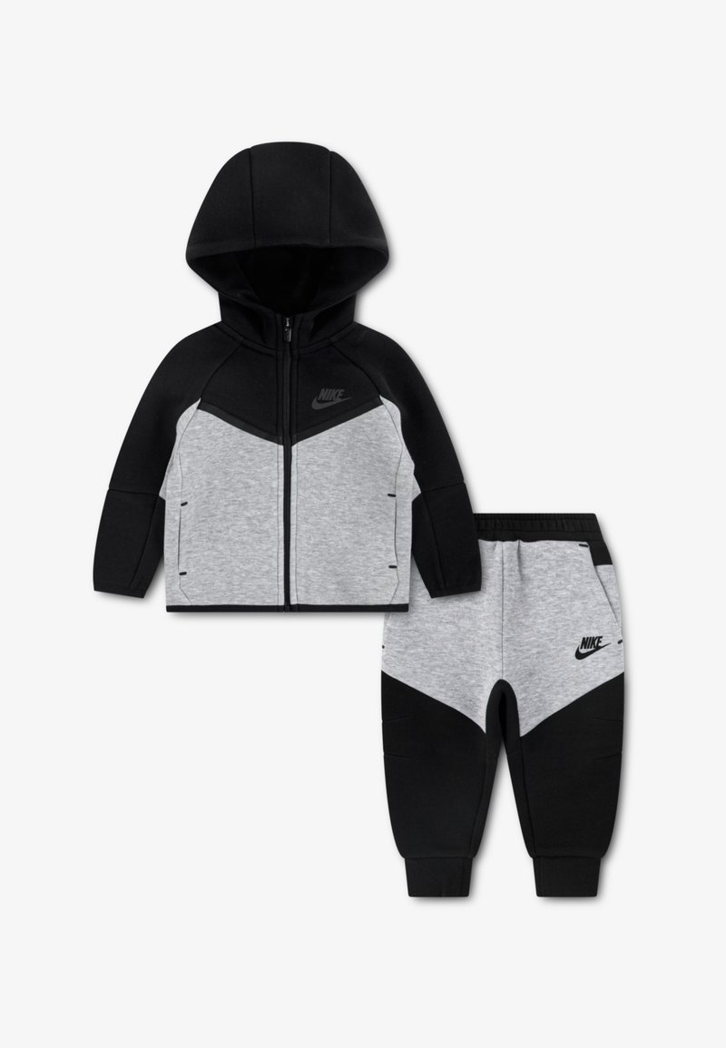 Διχρωμία φόρμας δύο τεμαχίων της Nike με ζακέτα φερμουάρ με κουκούλα σε μαύρο και γκρι με σχέδιο με βέλη, συνδυασμένη με αντίστοιχο στενό παντελόνι φόρμας.