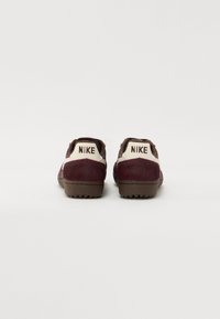 Nike batai su rudais ir burgundiškais kailio tipo raštais, smėlio spalvos akcentais ir guminėmis padangomis. Ant kulnų matoma "NIKE" prekės ženklas.