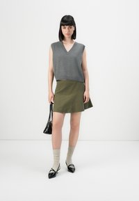 Young woman with black bob haircut wearing sleeveless gray sweater, olive green skirt, beige socks, and black shoes, holding a black handbag.