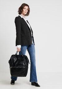 Mochila de cuero negro con un bolsillo frontal con cremallera, diseño elegante, dos asas y superficie texturizada, llevada por una persona con un blazer y jeans.
