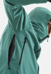 Person wearing teal hooded jacket with multiple black zippered vents, raising arm over head against a plain background.